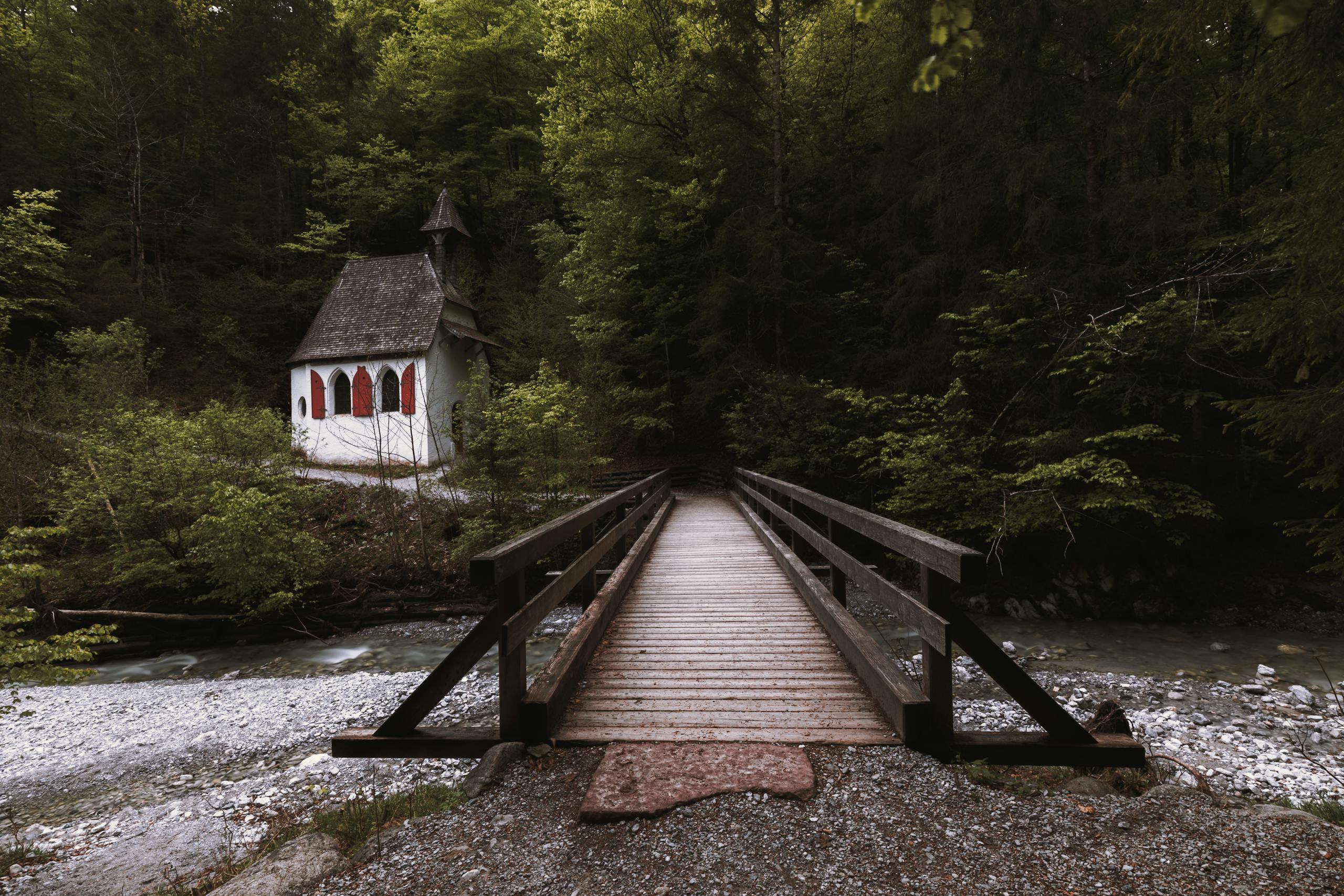 Idyllic scene of a small chapel in a lush forest by a wooden bridge and creek.